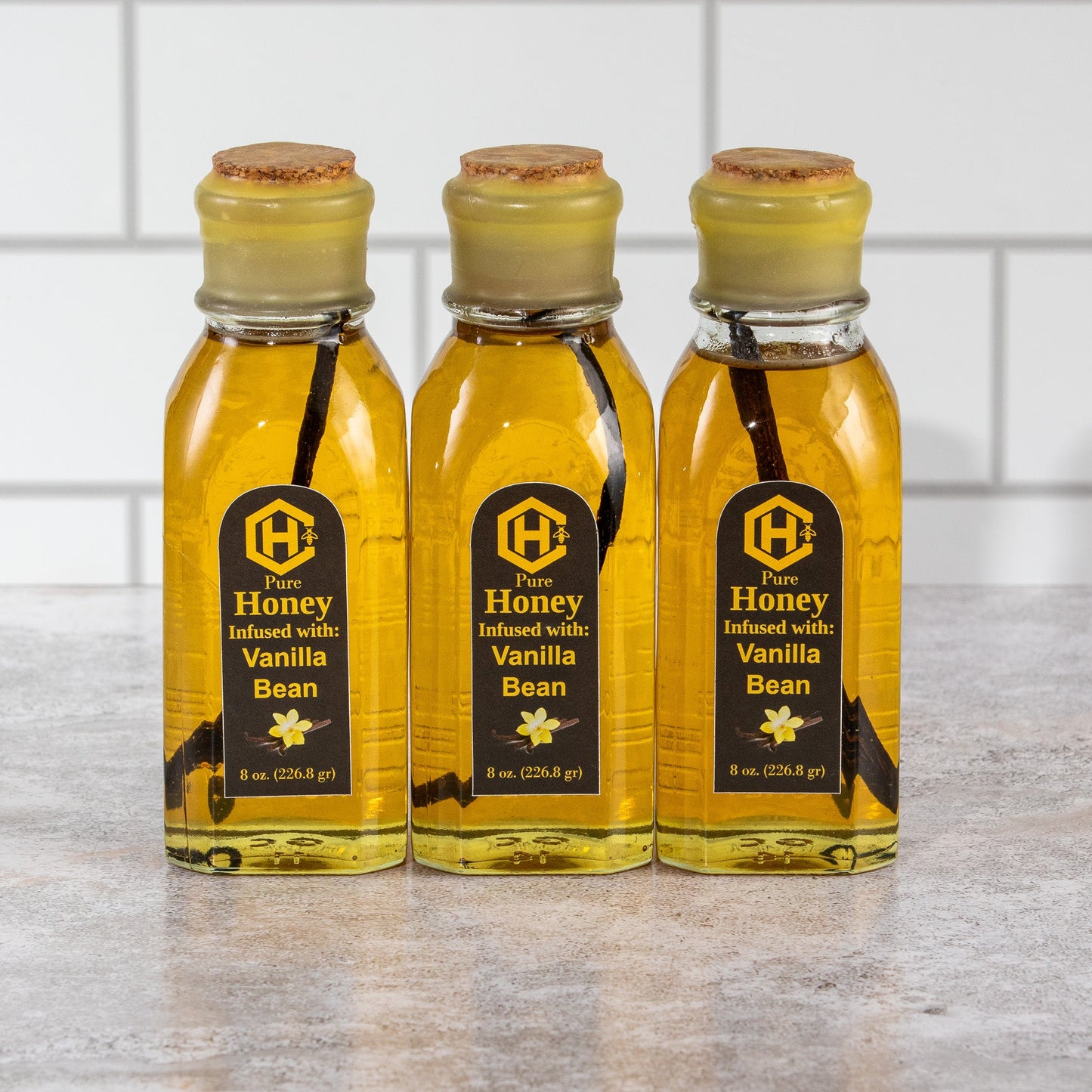 Three bottles of Honey Vanilla Bean syrup on a marble countertop with a tiled wall background.